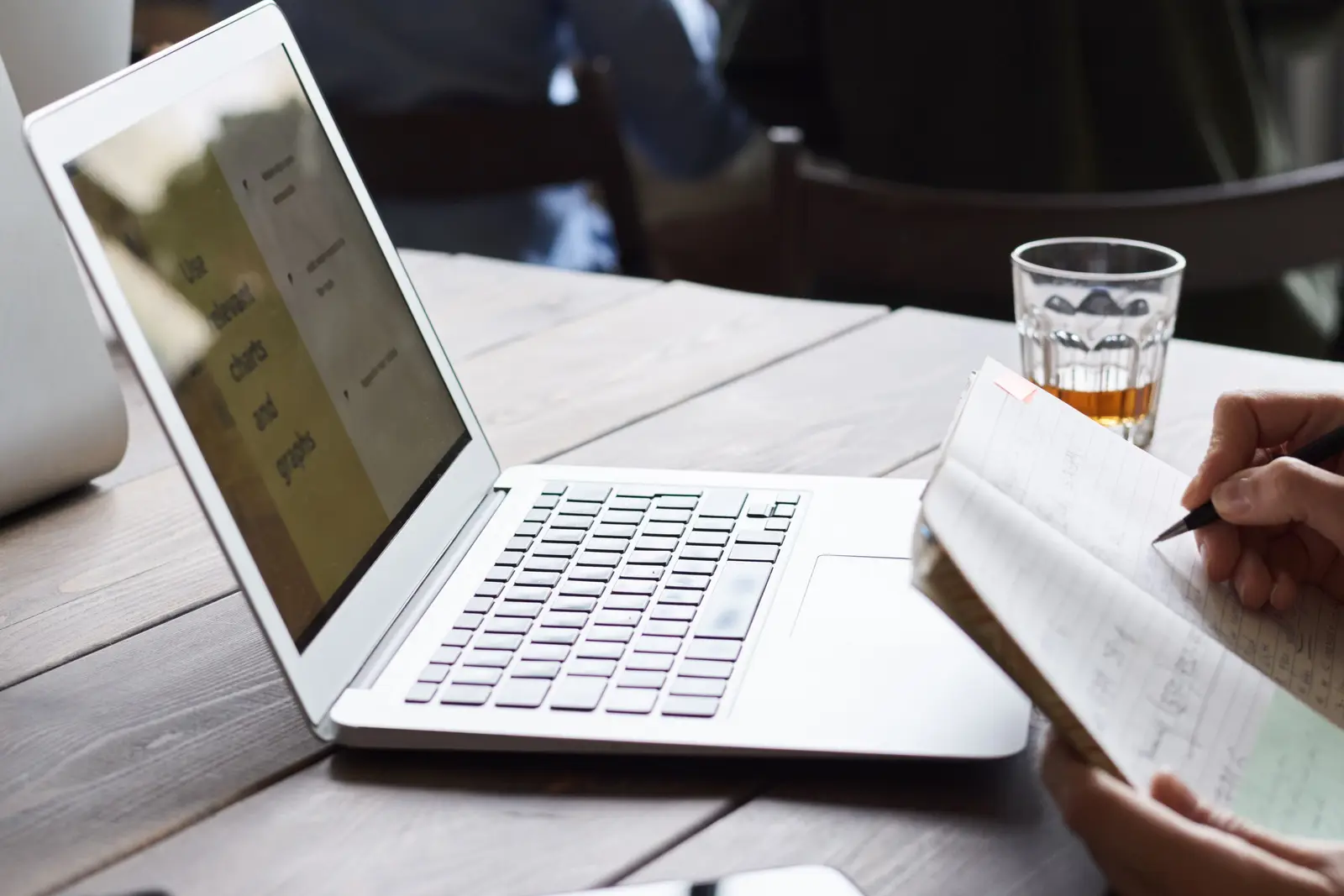 Ordinateur portable ouvert sur une table de réunion pendant qu'une personne prend des notes, équipe au travail en arrière-plan