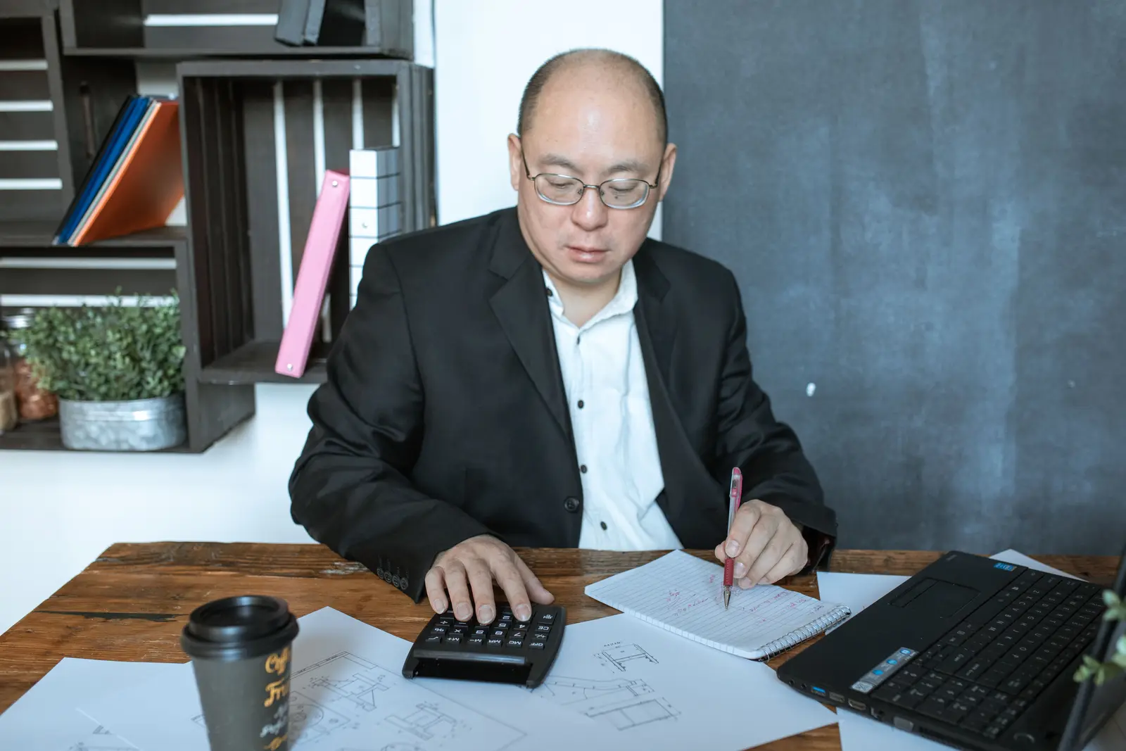 Dirigeant assis à son bureau, calculatrice dans une main et stylo dans l'autre, en train de vérifier des documents à côté de son ordinateur portable